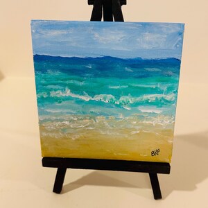 May include: A small square canvas painting of a beach scene featuring a yellow sandy shore, turquoise and blue ocean waves, and a light blue sky, displayed on a black wooden tripod easel. The artist's initials BKF are visible in the bottom right.