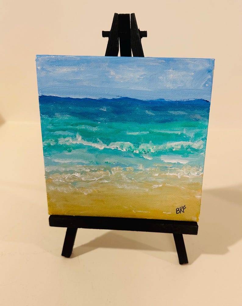 May include: A small square canvas painting of a beach scene featuring a yellow sandy shore, turquoise and blue ocean waves, and a light blue sky, displayed on a black wooden tripod easel. The artist's initials BKF are visible in the bottom right.