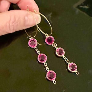May include: A pair of gold-toned hoop earrings featuring three dangling circular pink faceted stones on each earring. The stones are set in metal bezels and connected by small gold-toned links, creating a vertical drop design that hangs from the hoop.