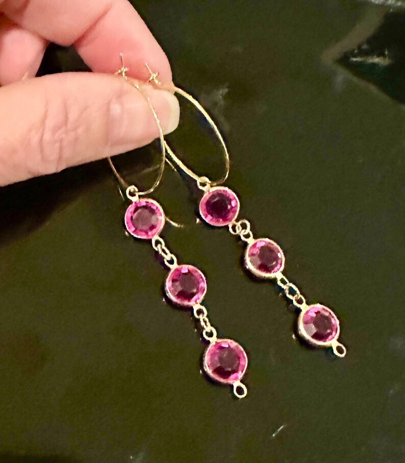 May include: A pair of gold-toned hoop earrings featuring three dangling circular pink faceted stones on each earring. The stones are set in metal bezels and connected by small gold-toned links, creating a vertical drop design that hangs from the hoop.