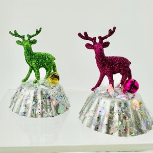 May include: Two decorative ornaments featuring glittery reindeer figures, one green and one magenta, standing atop silver fluted bases covered in iridescent confetti. Each base is accented with a small metallic jingle bell in gold or pink.