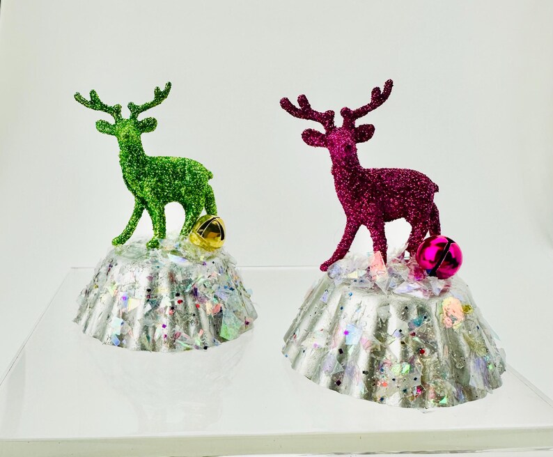 May include: Two decorative ornaments featuring glittery reindeer figures, one green and one magenta, standing atop silver fluted bases covered in iridescent confetti. Each base is accented with a small metallic jingle bell in gold or pink.