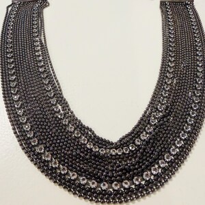 May include: A multi-strand bib necklace featuring dark gunmetal-toned ball chains arranged in a wide, curved silhouette. The design incorporates two distinct rows of clear, round-cut rhinestones set within the dark metal chain structure.