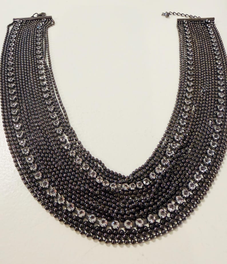 May include: A multi-strand bib necklace featuring dark gunmetal-toned ball chains arranged in a wide, curved silhouette. The design incorporates two distinct rows of clear, round-cut rhinestones set within the dark metal chain structure.