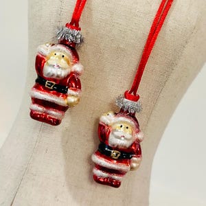 May include: Two small Santa Claus ornaments made of glass, each featuring a red suit, black belt with a gold buckle, and white trim. The figures have silver tinsel accents at the top and are suspended by thin red fabric cords for hanging.