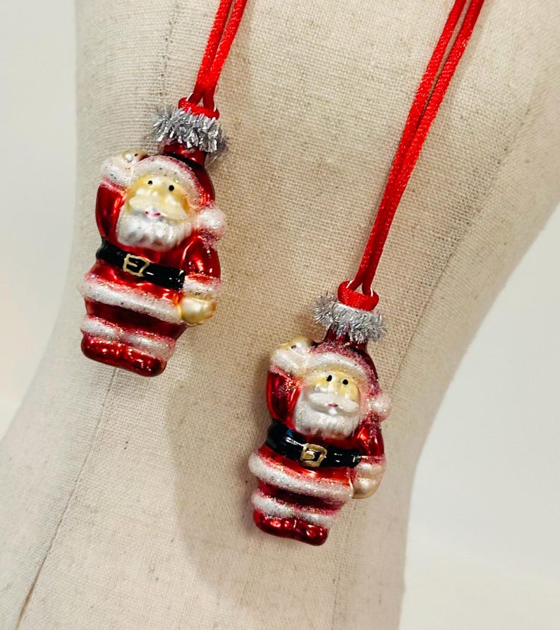 May include: Two small Santa Claus ornaments made of glass, each featuring a red suit, black belt with a gold buckle, and white trim. The figures have silver tinsel accents at the top and are suspended by thin red fabric cords for hanging.