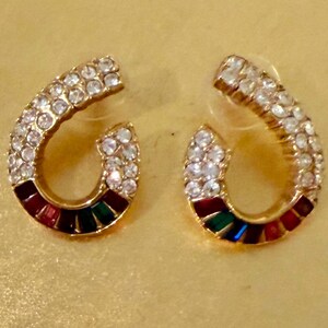 May include: A pair of horseshoe-shaped stud earrings in a gold-tone metal finish. The upper portion of each earring is encrusted with clear white crystals, while the lower curved section features a row of alternating red, green, and blue stones.