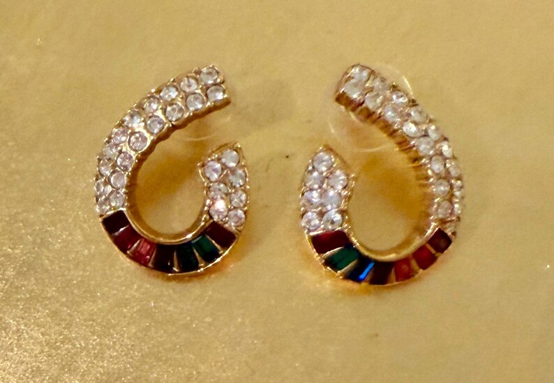 May include: A pair of horseshoe-shaped stud earrings in a gold-tone metal finish. The upper portion of each earring is encrusted with clear white crystals, while the lower curved section features a row of alternating red, green, and blue stones.