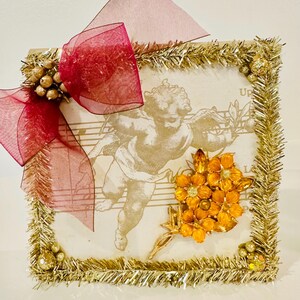 May include: A square decorative plaque featuring a sepia-toned cherub illustration over musical notation. The border is trimmed with gold tinsel and small gold spheres. A large, sheer burgundy ribbon bow is attached to the top left corner.