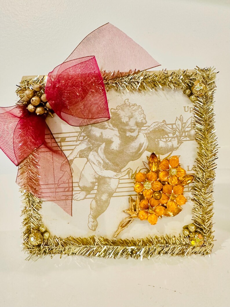 May include: A square decorative plaque featuring a sepia-toned cherub illustration over musical notation. The border is trimmed with gold tinsel and small gold spheres. A large, sheer burgundy ribbon bow is attached to the top left corner.