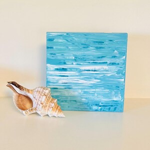 May include: A square canvas painting featuring abstract horizontal brushstrokes in shades of light blue, turquoise, and white, resembling ocean waves. Beside the painting sits a spiral-shaped seashell with tan and brown striped patterns.