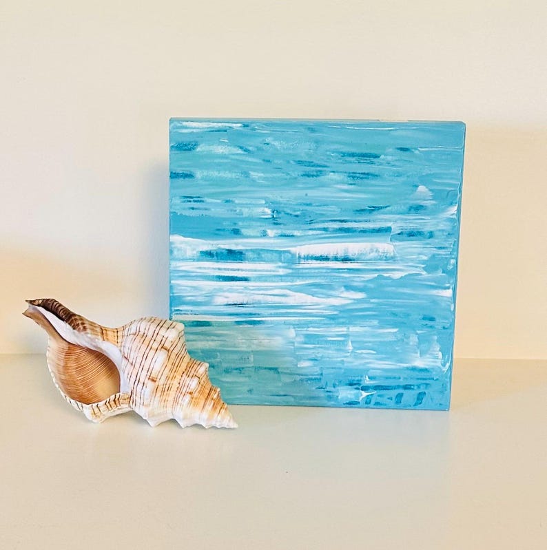 May include: A square canvas painting featuring abstract horizontal brushstrokes in shades of light blue, turquoise, and white, resembling ocean waves. Beside the painting sits a spiral-shaped seashell with tan and brown striped patterns.