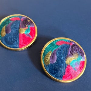 May include: A pair of round earrings featuring a gold-tone metal rim and a swirling marbled enamel inlay. The colorful surface displays a vibrant mix of deep blue, bright pink, purple, teal, and shimmering gold paint in a fluid, abstract pattern.