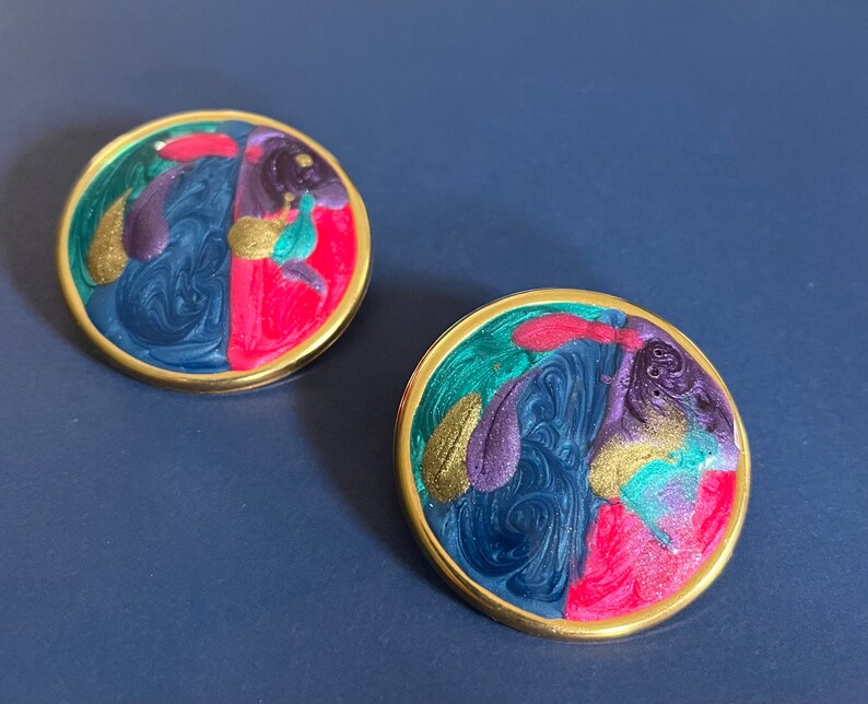 May include: A pair of round earrings featuring a gold-tone metal rim and a swirling marbled enamel inlay. The colorful surface displays a vibrant mix of deep blue, bright pink, purple, teal, and shimmering gold paint in a fluid, abstract pattern.