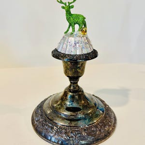 May include: A tarnished silver-toned metal candlestick base topped with a white, glittery, fluted cupcake liner. A small, bright green glittery deer figurine stands on the liner next to a single, small, gold-toned jingle bell ornament.