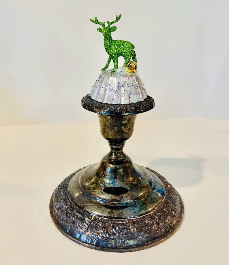 May include: A tarnished silver-toned metal candlestick base topped with a white, glittery, fluted cupcake liner. A small, bright green glittery deer figurine stands on the liner next to a single, small, gold-toned jingle bell ornament.