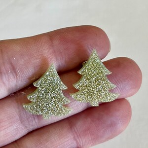 May include: A pair of stud earrings shaped like Christmas trees. Each earring features a flat, triangular silhouette with tiered branches and a small trunk at the base. The surface of the earrings is covered in a fine, light gold glitter texture.