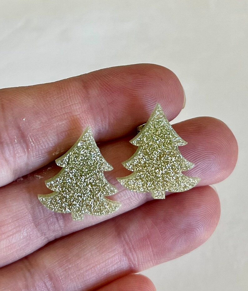 May include: A pair of stud earrings shaped like Christmas trees. Each earring features a flat, triangular silhouette with tiered branches and a small trunk at the base. The surface of the earrings is covered in a fine, light gold glitter texture.