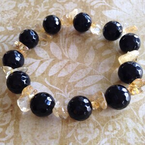 May include: A stretch bracelet featuring alternating large, round, faceted black beads and smaller, irregular, translucent yellow-gold crystal chips. The black beads have a glossy, reflective surface, while the yellow-gold chips provide a contrasting texture and color throughout the circular arrangement.