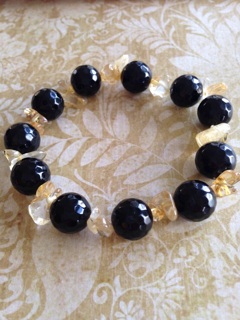 May include: A stretch bracelet featuring alternating large, round, faceted black beads and smaller, irregular, translucent yellow-gold crystal chips. The black beads have a glossy, reflective surface, while the yellow-gold chips provide a contrasting texture and color throughout the circular arrangement.