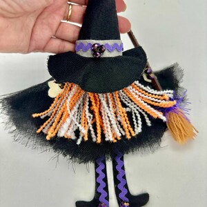 May include: A hanging witch ornament featuring a black pointed hat with a grey band and purple trim, a black tulle skirt, and a yarn beard in orange, white, and black. The figure has dangling legs with purple wavy ribbon and a small broom accessory.