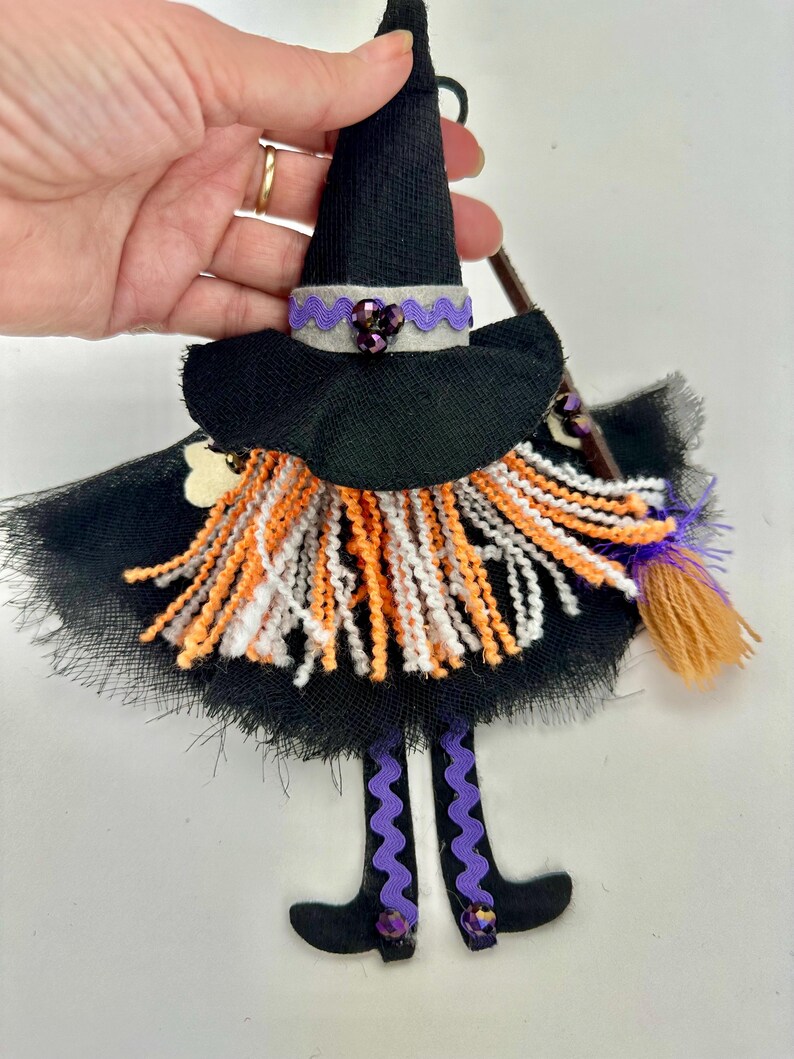 May include: A hanging witch ornament featuring a black pointed hat with a grey band and purple trim, a black tulle skirt, and a yarn beard in orange, white, and black. The figure has dangling legs with purple wavy ribbon and a small broom accessory.