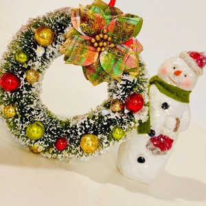 May include: A miniature Christmas wreath featuring green pine needles with white snow dusting, decorated with small red, gold, and lime green ornaments, topped with a plaid poinsettia, positioned next to a white snowman figurine wearing a red hat and green scarf.