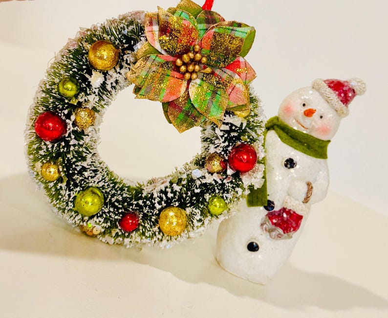 May include: A miniature Christmas wreath featuring green pine needles with white snow dusting, decorated with small red, gold, and lime green ornaments, topped with a plaid poinsettia, positioned next to a white snowman figurine wearing a red hat and green scarf.