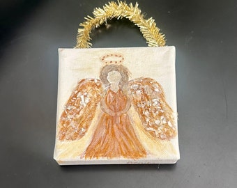 May include: A square canvas painting of an angel with a brown dress and textured gold wings, topped with a gold tinsel loop for hanging. The angel features a simple halo and is rendered in shades of brown, tan, and metallic gold on a light background.
