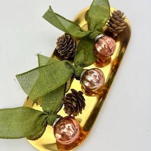 May include: A gold-colored, oblong tray holds three decorative ornaments. Each ornament is a small, round, copper-colored ball tied with a green ribbon and accompanied by a small pinecone. The arrangement is set against a white background.