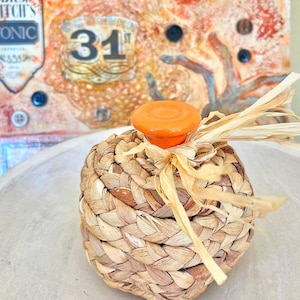 May include: A woven basket with an orange knob on top, tied with raffia. The basket is made of natural fibers and has a round shape. The background features a vintage-style artwork with the number 31.