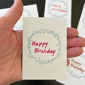 May include: A small rectangular white card featuring a hand-drawn light blue circular wreath design with the text Happy Birthday written in pink ink in the center. Other similar cards with different text are visible in the background.
