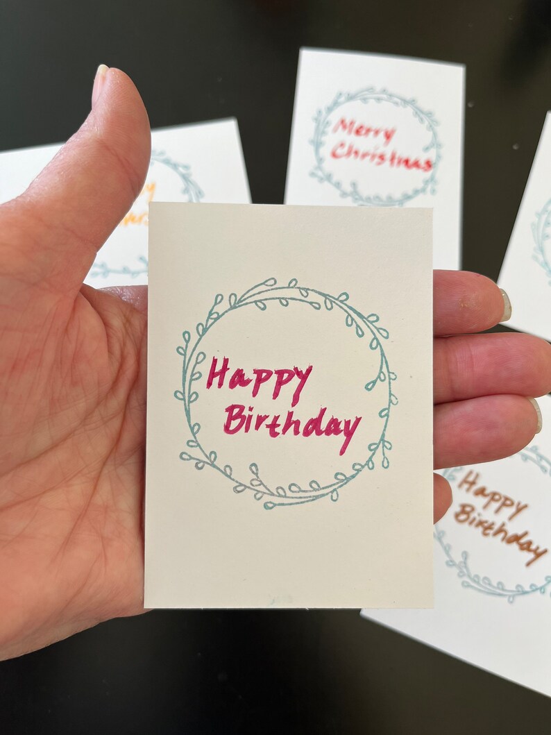 May include: A small rectangular white card featuring a hand-drawn light blue circular wreath design with the text Happy Birthday written in pink ink in the center. Other similar cards with different text are visible in the background.