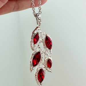 May include: Silver-toned pendant necklace featuring a leaf design with five marquise-cut, deep red gemstones. The pendant is encrusted with small, clear crystals, and hangs from a delicate chain. The necklace is held up against a light background.