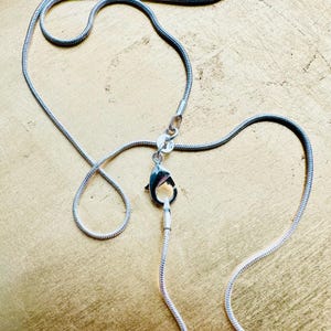 May include: A silver-toned snake chain necklace featuring a polished finish and a secure lobster claw clasp. The flexible, rounded chain links create a smooth, continuous appearance, with the metal clasp positioned centrally to connect the ends.