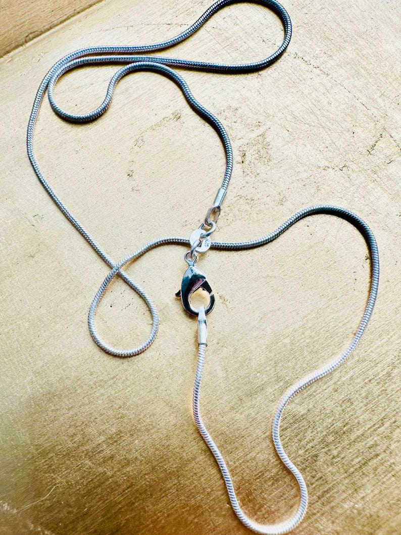 May include: A silver-toned snake chain necklace featuring a polished finish and a secure lobster claw clasp. The flexible, rounded chain links create a smooth, continuous appearance, with the metal clasp positioned centrally to connect the ends.