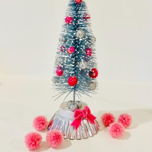 May include: A miniature blue bottle brush tree with white snow-like flocking, decorated with small shiny red and silver spherical ornaments. The tree stands on a silver fluted base with a pink ribbon bow, surrounded by several small pink pom-poms.