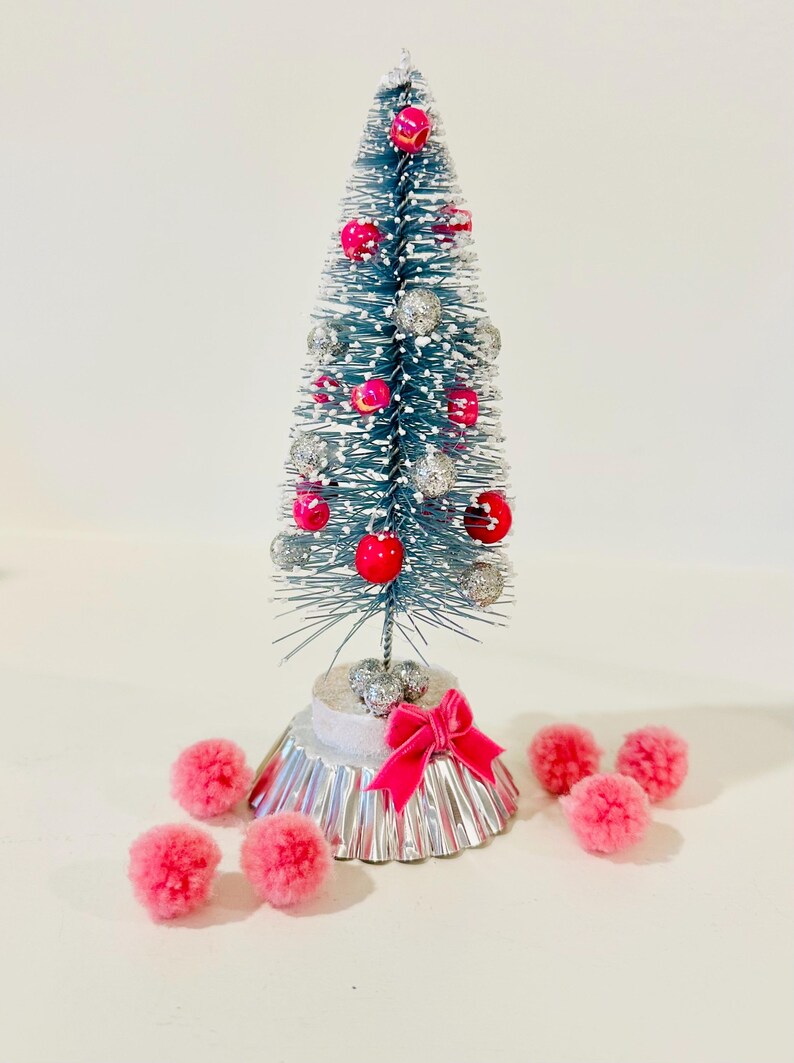 May include: A miniature blue bottle brush tree with white snow-like flocking, decorated with small shiny red and silver spherical ornaments. The tree stands on a silver fluted base with a pink ribbon bow, surrounded by several small pink pom-poms.