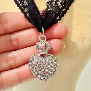 May include: A black lace choker necklace featuring a silver-toned pendant shaped like a heart topped with a small crown. The heart pendant is encrusted with numerous clear, sparkling rhinestones that cover its entire front surface.