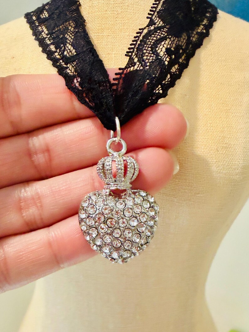 May include: A black lace choker necklace featuring a silver-toned pendant shaped like a heart topped with a small crown. The heart pendant is encrusted with numerous clear, sparkling rhinestones that cover its entire front surface.