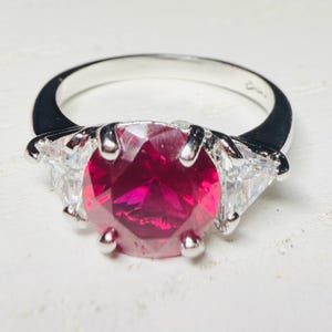 May include: A silver ring featuring a large, round, faceted deep red gemstone at the center, held by four prongs. On each side of the central stone, there is a smaller, triangular, clear faceted gemstone set into the polished silver band.