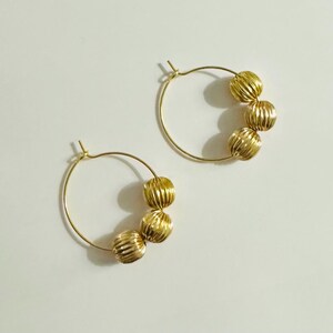 May include: A pair of gold hoop earrings, each featuring three small, round, vertically ribbed beads threaded onto the thin circular wire. The metal has a polished, reflective finish, and the hoops are designed with a simple post-and-clasp closure.