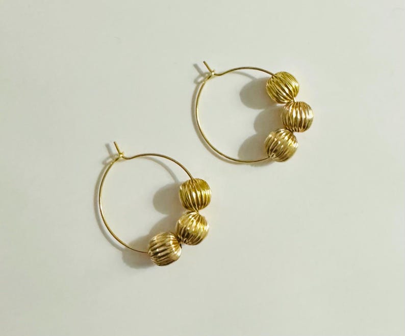 May include: A pair of gold hoop earrings, each featuring three small, round, vertically ribbed beads threaded onto the thin circular wire. The metal has a polished, reflective finish, and the hoops are designed with a simple post-and-clasp closure.