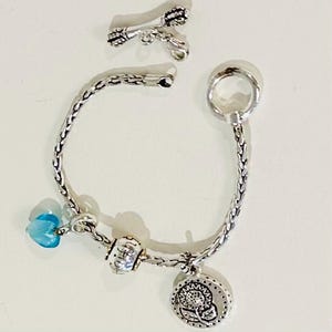 May include: A silver-toned chain bracelet featuring a circular toggle clasp and three charms: a translucent light blue heart, a small textured bead, and a round pendant embossed with a sunflower design, accompanied by a separate silver arrow charm.