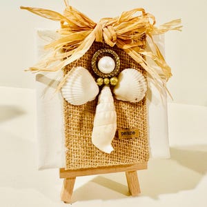 May include: A small white canvas on a wooden easel featuring a burlap strip decorated with a shell angel. The angel has a pearl head, gold ring halo, two white scallop shell wings, and a long shell body. A raffia bow sits on top with a small tag reading peace.
