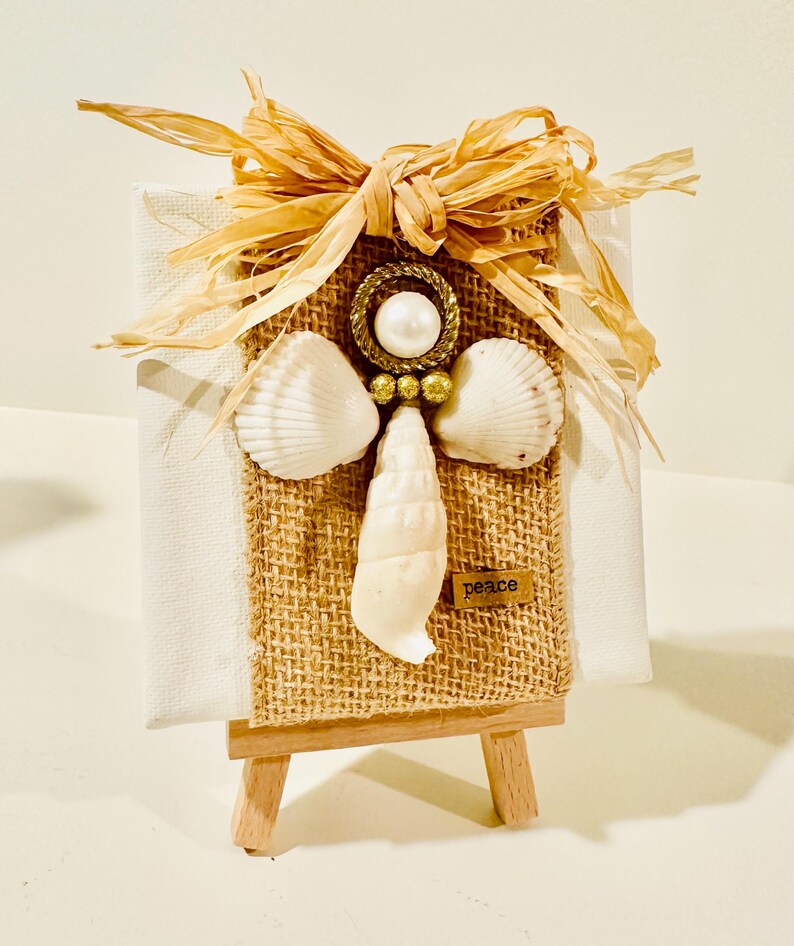 May include: A small white canvas on a wooden easel featuring a burlap strip decorated with a shell angel. The angel has a pearl head, gold ring halo, two white scallop shell wings, and a long shell body. A raffia bow sits on top with a small tag reading peace.