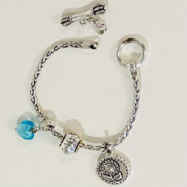 May include: A silver-toned chain bracelet featuring a toggle clasp, a translucent blue heart-shaped charm, a small round patterned bead, and a circular charm embossed with a sunflower design, accompanied by a separate silver-toned arrow-shaped charm.