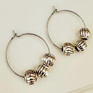 May include: A pair of silver-toned hoop earrings, each featuring three small, round, vertically ribbed beads threaded onto the thin wire hoop. The metal has a polished, reflective finish, and the hoops are circular with a simple clasp mechanism.