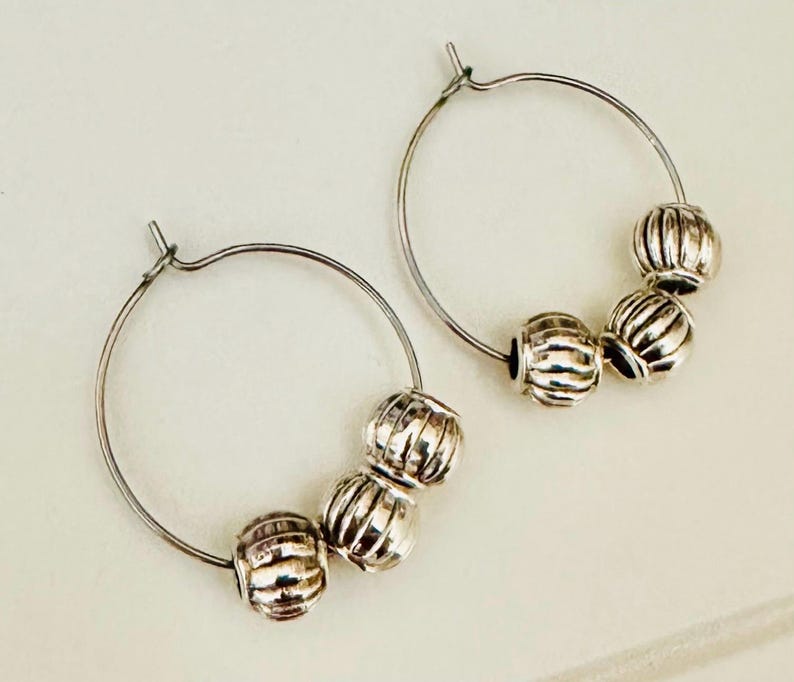 May include: A pair of silver-toned hoop earrings, each featuring three small, round, vertically ribbed beads threaded onto the thin wire hoop. The metal has a polished, reflective finish, and the hoops are circular with a simple clasp mechanism.