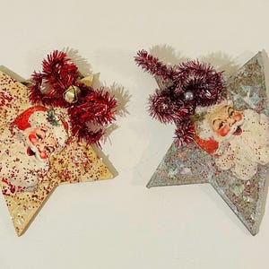 May include: Two star-shaped ornaments featuring vintage-style Santa Claus illustrations. The left star is cream with red glitter, while the right star is silver with iridescent glitter. Both are topped with red tinsel bows and small bells.